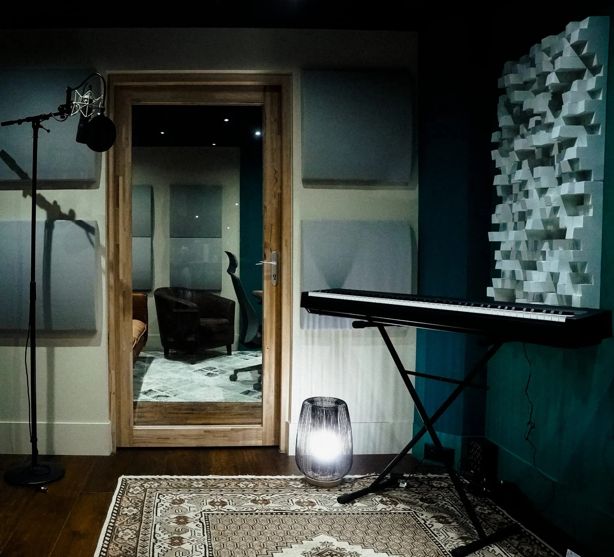 Cabine d’enregistrement avec micro sur pied, clavier et diffuseurs acoustiques, vue vers la régie